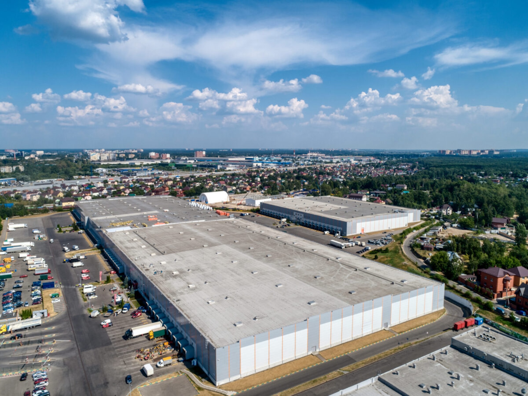 Huge warehouse building with flat roof and cars on parking site under blue sky with fluffy clouds near city in summer bird eye view