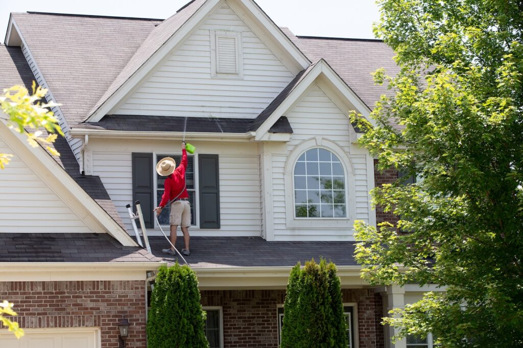 Homeowner washing the exterior of his house in spring balancing on the lower roof to spray the second floor facade with a handheld pressure sprayer
