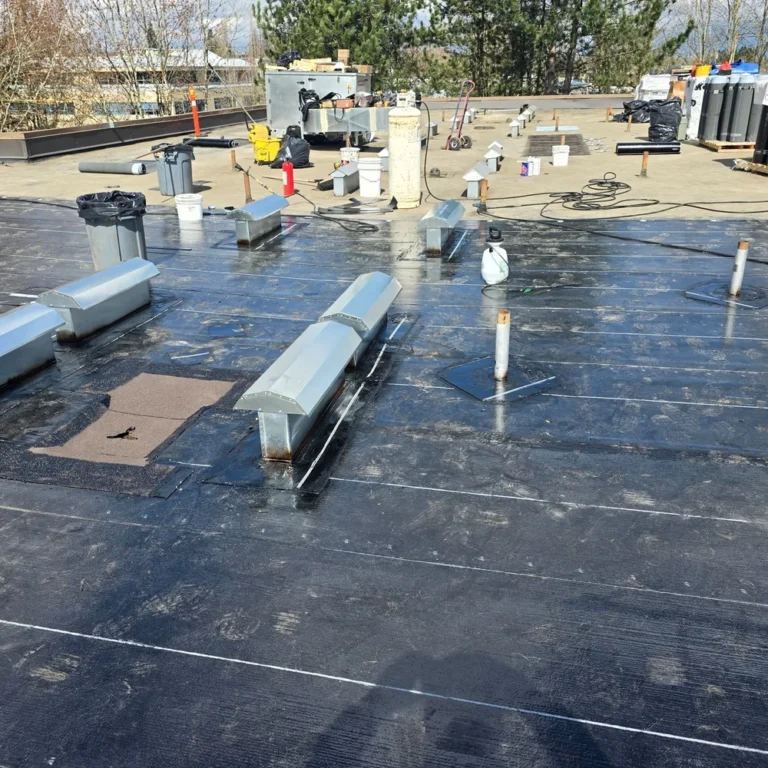 Flat commercial roof with black membrane, metal vents and pipes, tools and materials scattered during roofing work