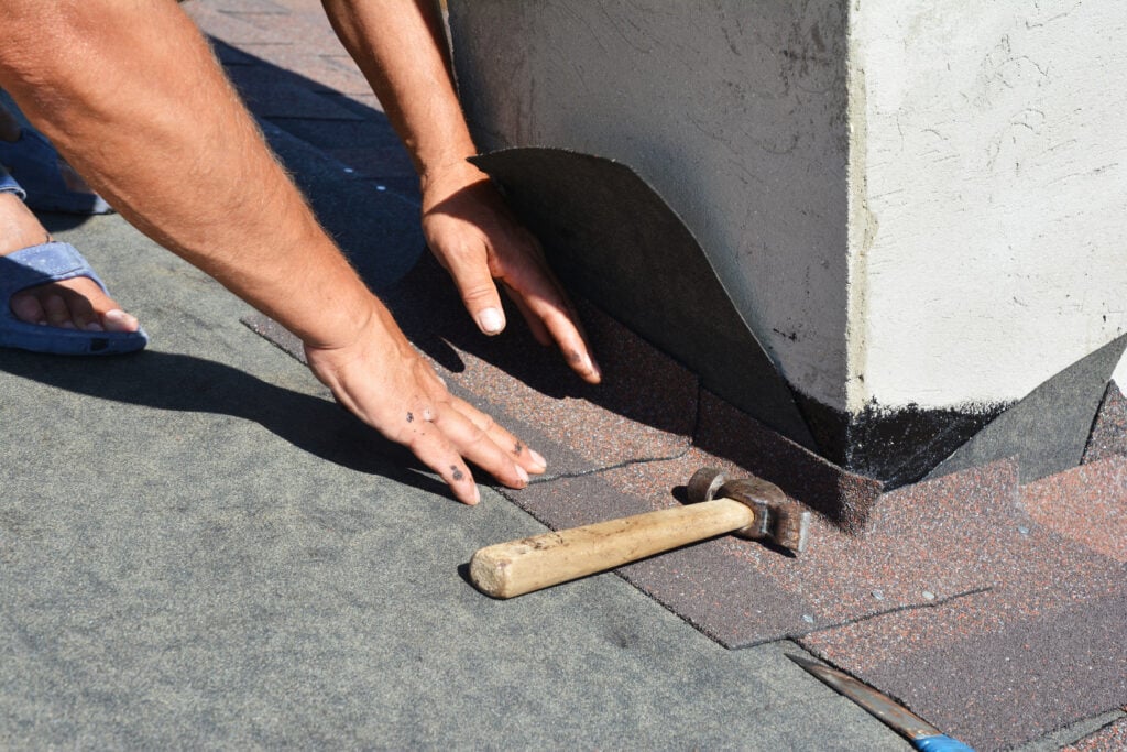 Asphalt shingles roofing construction. Roofer repair and laying asphalt shingles with vapor barrier in chimney problem area. Chimney waterproofing on house rooftop.
